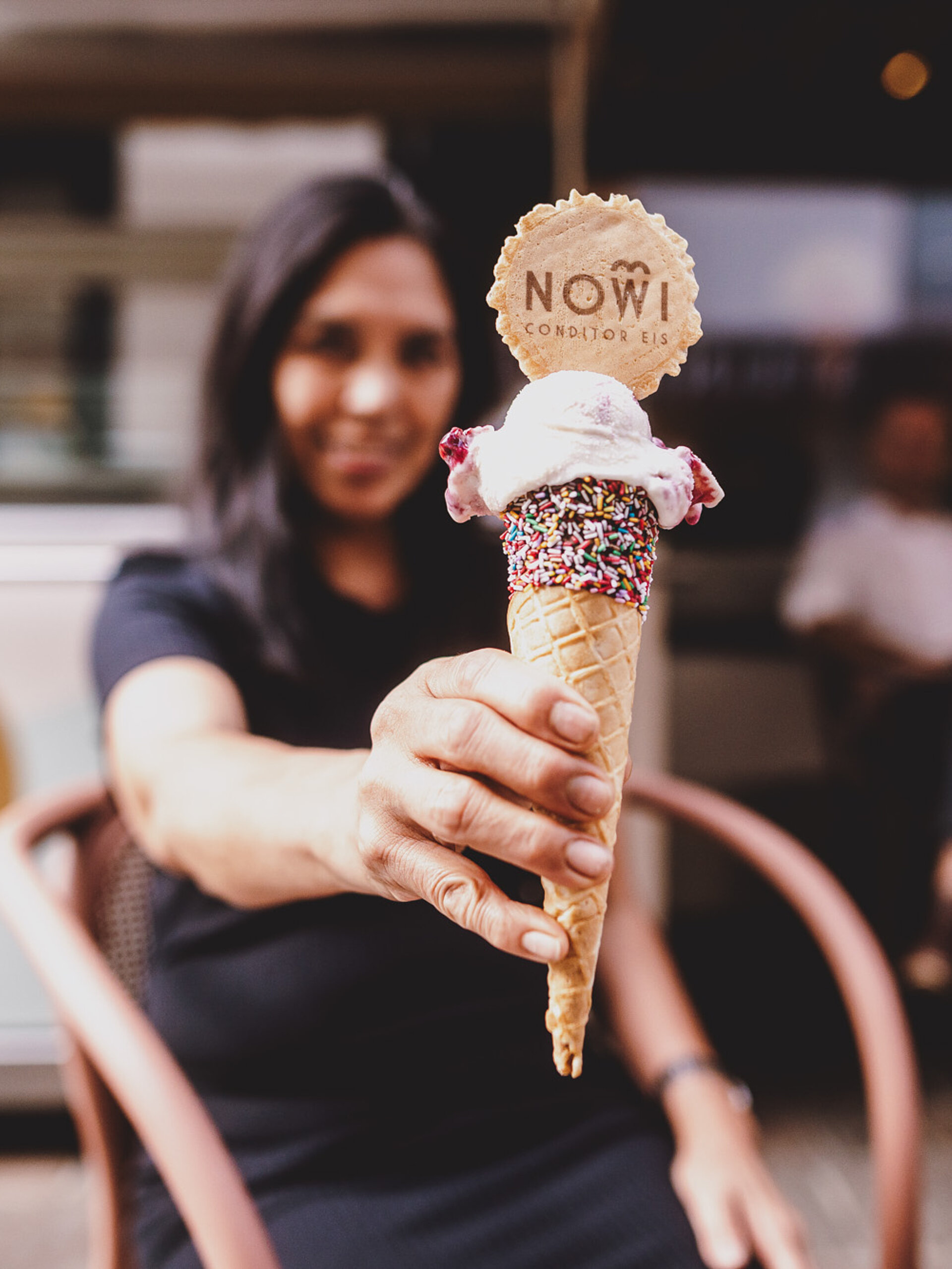 NOWI Eis Frau hält ein Eis in einer Waffel mit Streuseln und einer dekorativen Logo-Waffel. Im Hintergrund das Café Seegasse und Gäste, die den Außenbereich nutzen.