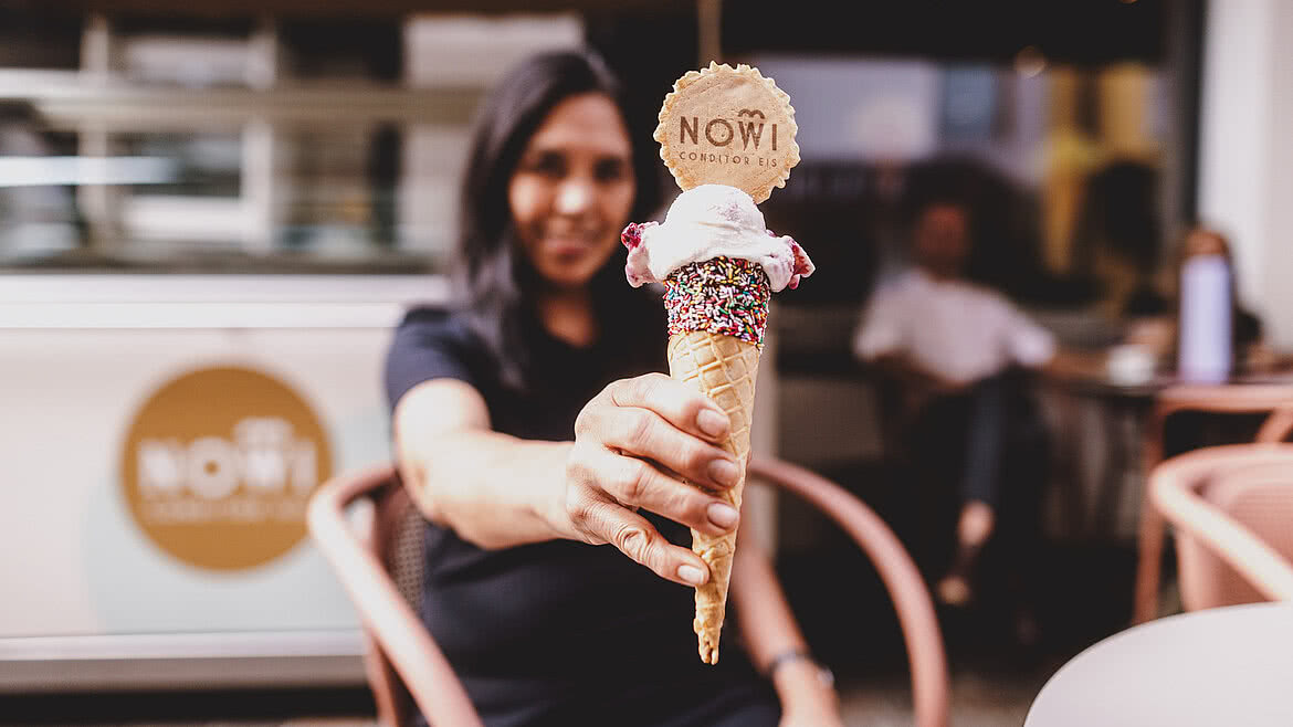 Auhof Hotel GmbH Frau hält ein Eis in einer Waffel mit Streuseln und einer dekorativen Logo-Waffel. Im Hintergrund das Café Seegasse und Gäste, die den Außenbereich nutzen.