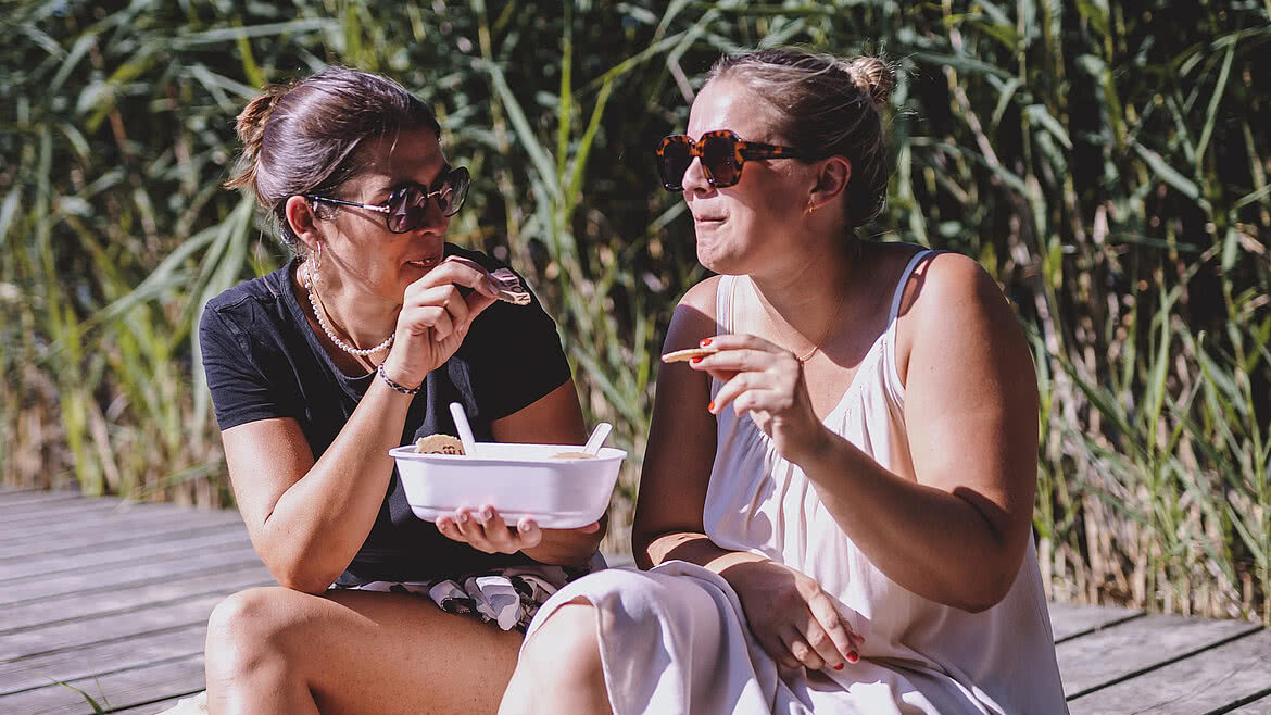 NOWI Eis Zwei Frauen, entspannt auf einem Holzsteg am Wasser sitzend, genießen Eis aus einer Schale, umgeben von hohen Gräsern im Hintergrund.