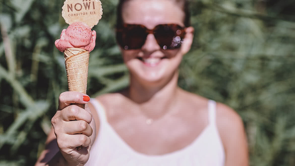 Frau im weißen Sommerkleid hält ein Eis mit einer NOWI-Dekorwaffel vor sich, grünes Schilf als unscharfer Hintergrund.