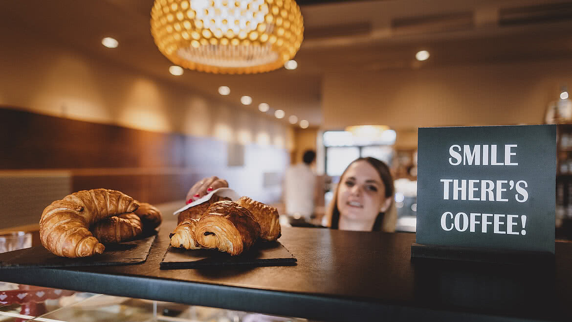 Auhof Hotel GmbH Frische Croissants auf einer schwarzen Schieferplatte in einem Café mit warmem Licht, im Hintergrund ein Schild mit der Aufschrift "Smile, there's coffee!" und eine freundliche Bedienung.