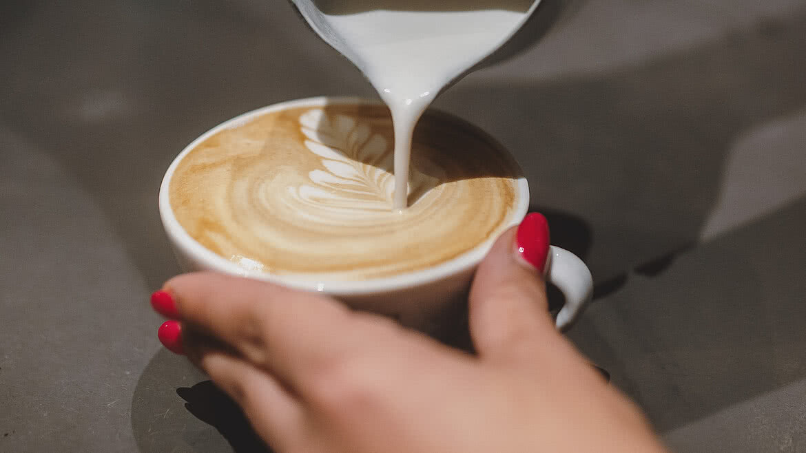 Auhof Hotel GmbH Detaillierte Ansicht von Latte Art, während Milch in eine Cappuccino-Tasse gegossen wird. Eine Hand hält die Tasse, die andere gießt mit einer Metallkanne.